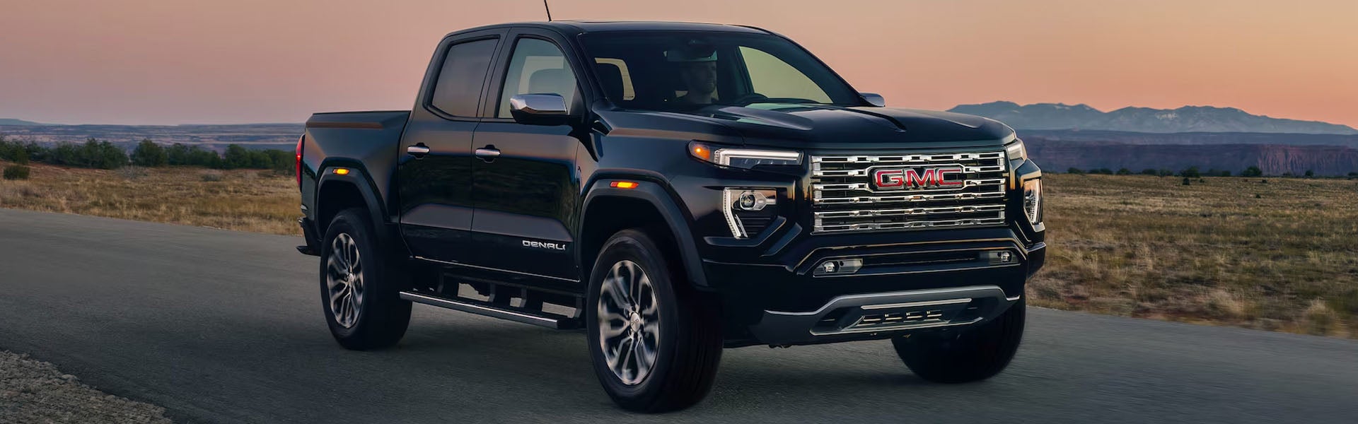 A black GMC Canyon pickup truck driving along a rural open road during sunset.