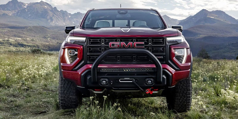 Front head-on view of a deep red GMC Canyon AT4X pickup truck parked in an open grassy field with mountains in the background.